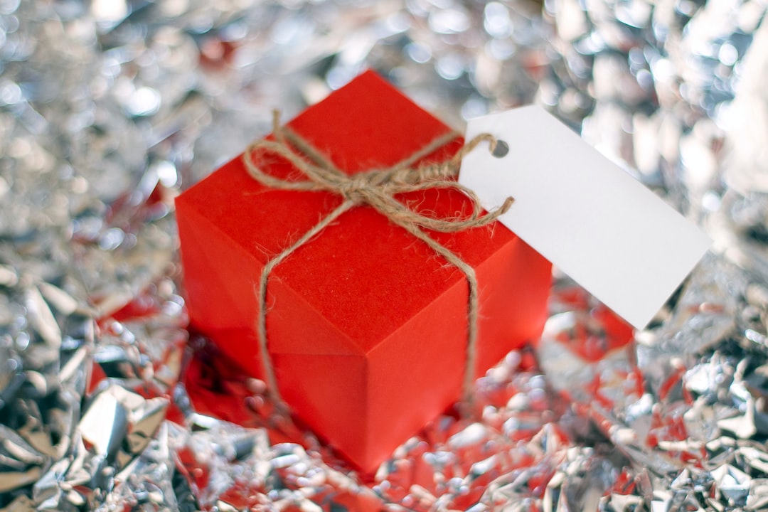 A small red gift box tied with twine and a blank tag sits on a surface covered in crumpled silver foil—an elegant choice for anniversary gifts to celebrate your love.