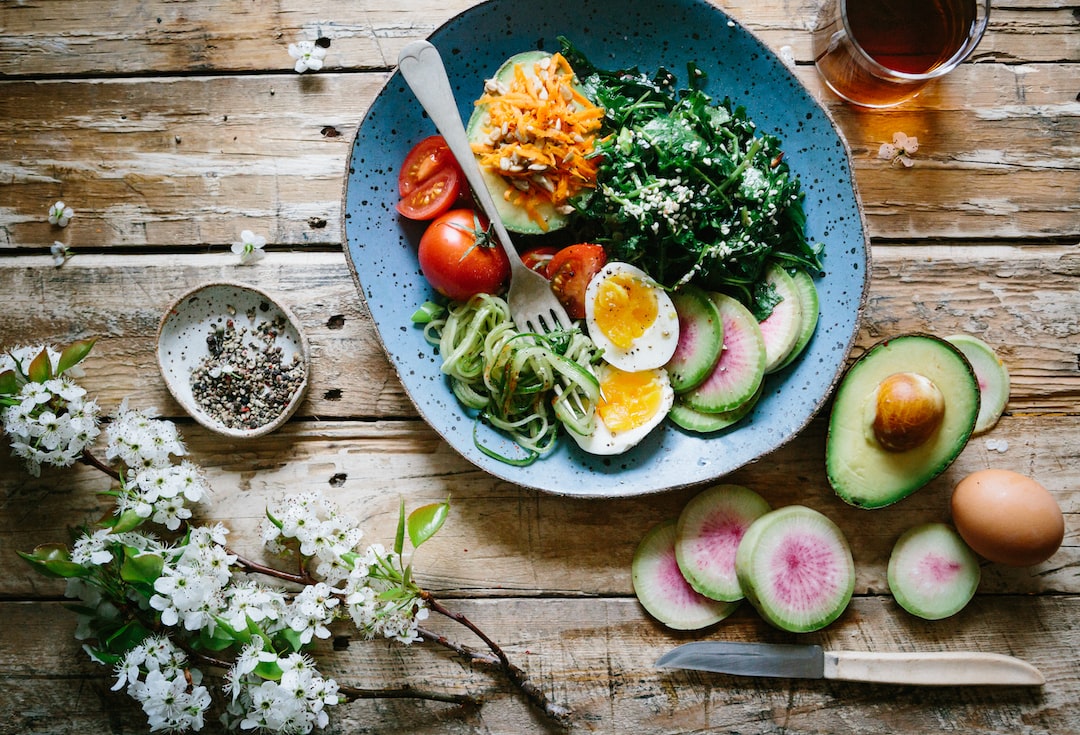 A blue plate holds sliced eggs, tomatoes, greens, shredded carrots, cucumber noodles, and avocado—perfect for a self-care meal. Sliced watermelon radish and half an avocado sit beside it on a rustic wooden table.