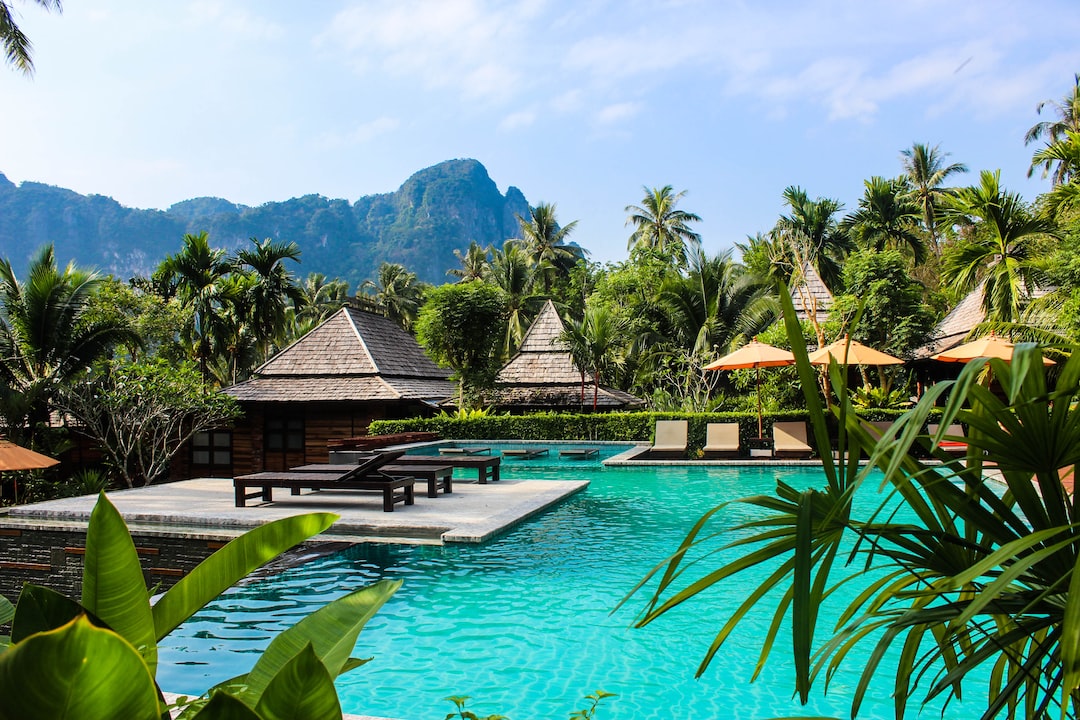 A tropical resort pool with wooden lounge chairs, surrounded by palm trees and mountains, under a clear blue sky—an ideal escape for those seeking relaxation at luxury eco-resorts.