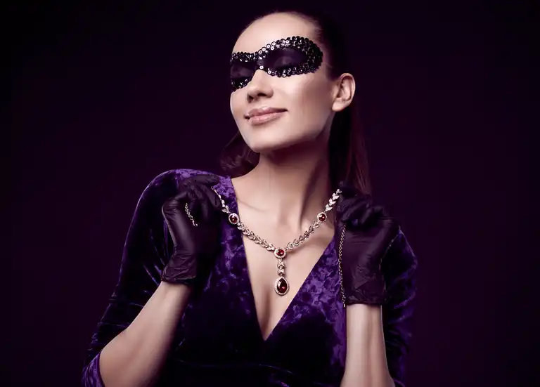 A woman wearing a black lace eye mask, black gloves, and a velvet purple dress displays a luxury gemstone necklace against a dark background.