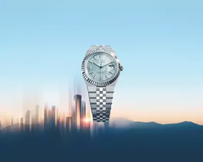A luxury silver wristwatch with a light blue face is centered above a blurred cityscape and mountains at sunrise.