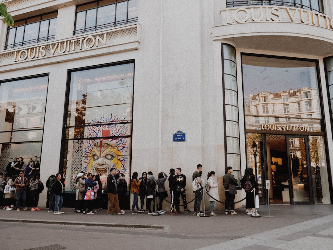People stand in a line outside a Louis Vuitton store on a city street in a fashion capital. The storefront displays large windows and the brand's name above the entrance, reflecting high-end shopping allure.