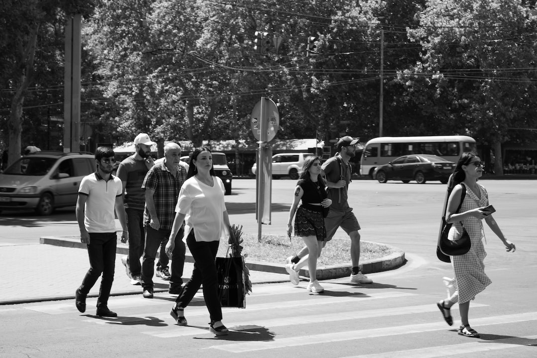 A group of people cross a city street at a crosswalk on a sunny day, moving with ultimate style as cars and trees line the background.