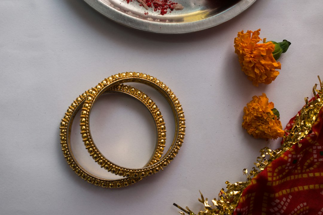 Two gold bangles add a touch of glamour on a white surface, paired with marigold flowers, a red-patterned cloth, and a metal plate with red powder.