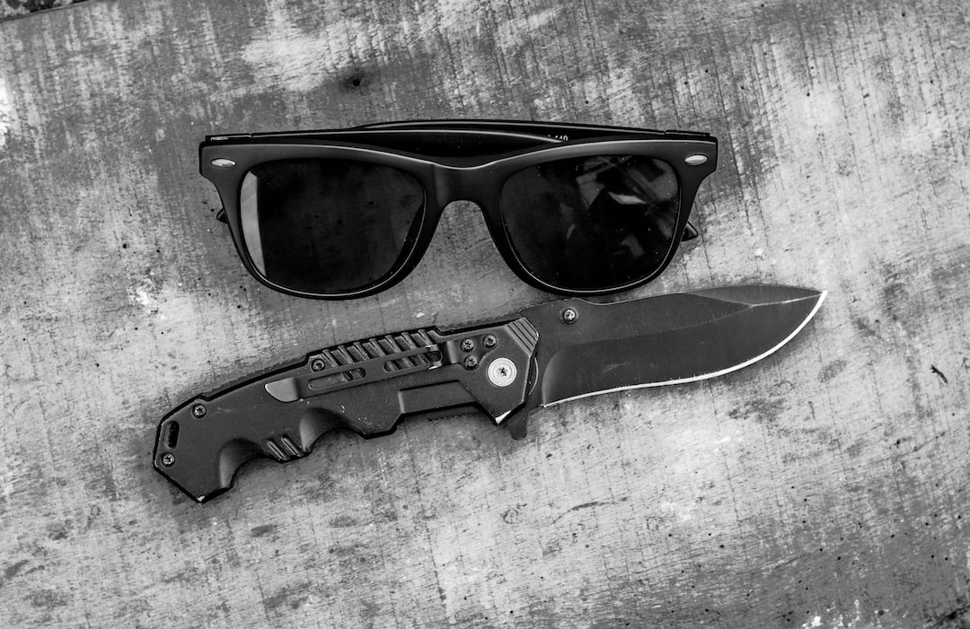 Black sunglasses and a folding knife with a black handle rest side by side on a weathered, textured surface in this black and white image, capturing an urban chic vibe with an edgy Alexander Wang-inspired aesthetic.