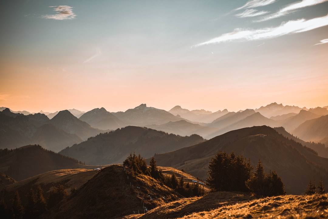 Mountain range with rolling hills and scattered trees, seen at sunset with a warm orange sky and layered silhouettes of distant peaks, radiating Alpine elegance reminiscent of Zurich's luxury mountain resorts.