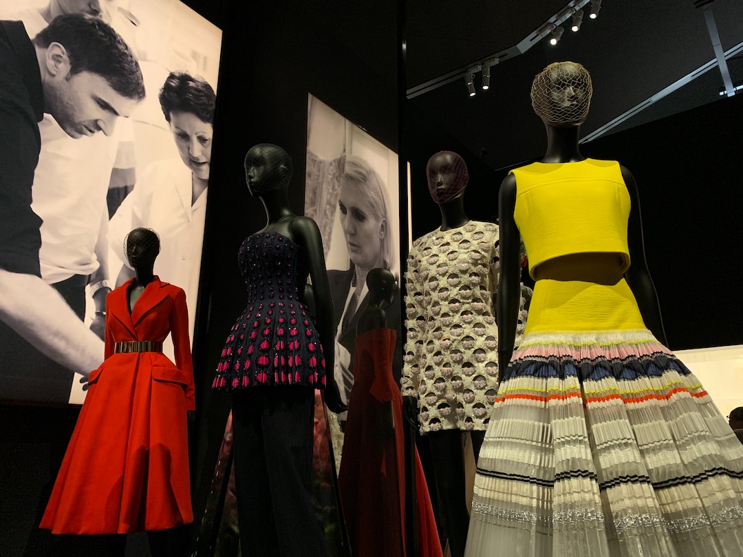 Four mannequins display colorful and patterned Armani designer dresses in a dimly lit exhibit, exuding modern elegance against a backdrop of large black-and-white fashion photos.