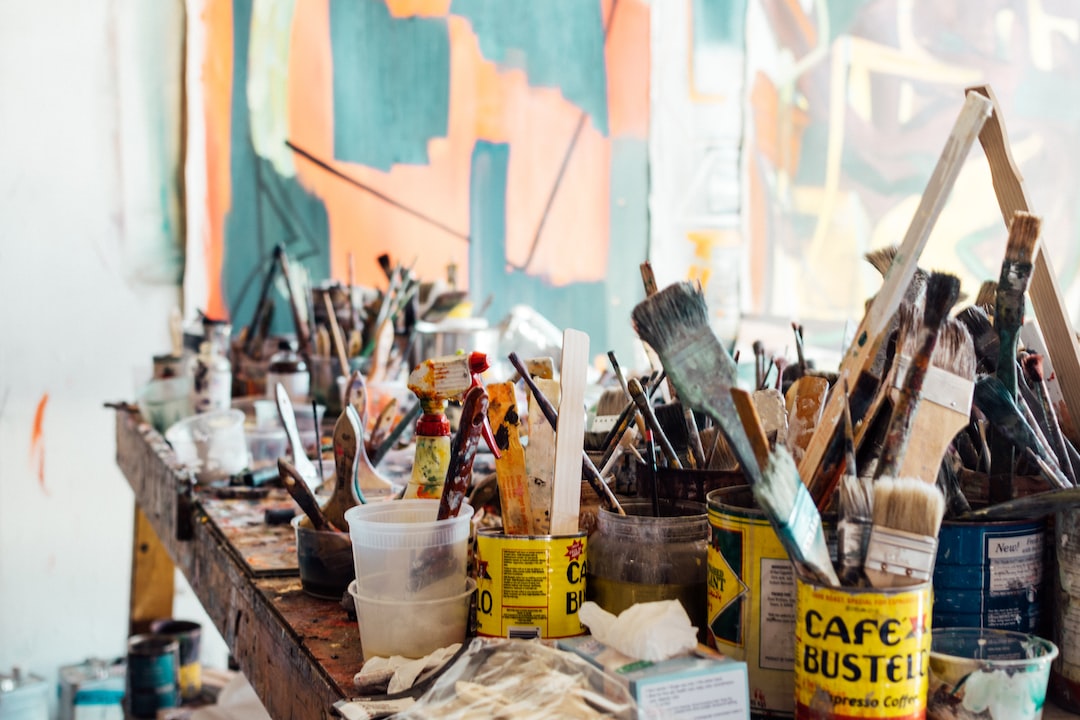 A cluttered artist's workspace with various paintbrushes, cans, jars, and art supplies on a wooden table in front of abstract paintings evokes the creative energy found in artistic Vienna and its inspiring Vienna art galleries.