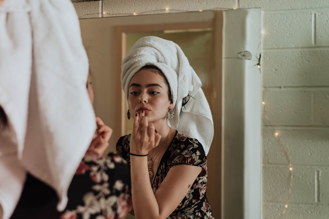 A woman with a towel wrapped around her head applies lipstick while looking at herself in a mirror, reflecting on expert advice and beauty tips tailored to her skin type.