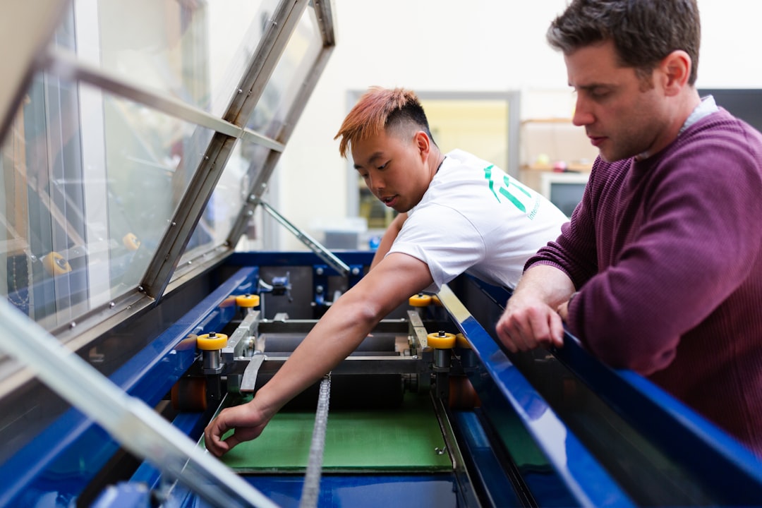 Two people operate and observe a piece of industrial machinery inside a workshop, with one adjusting controls and the other watching closely, much like an Anfänger learning eyebrow styling by observing experienced Augenbrauenstyling techniques.