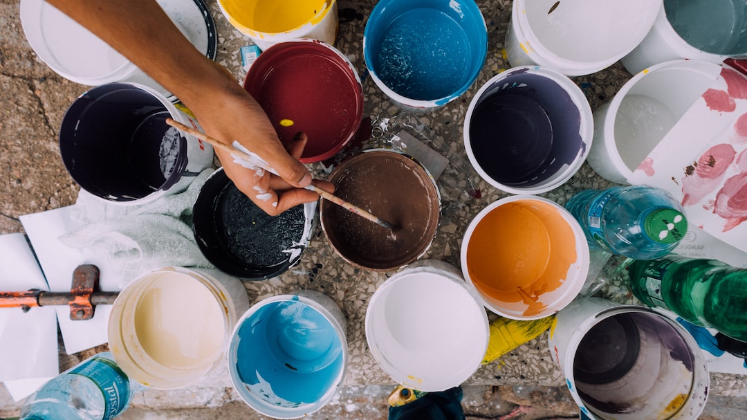 A person dips a paintbrush into a brown paint container surrounded by open buckets of various colored paints on the ground, capturing the creative spirit of the Berlin art scene.