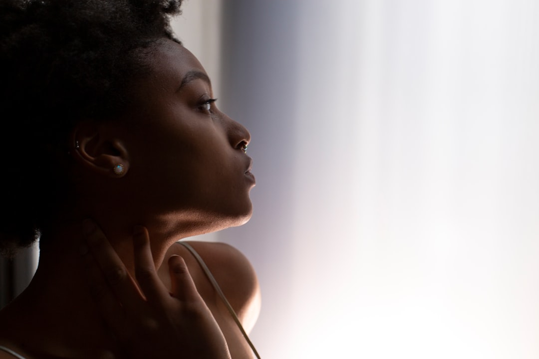 A person with short curly hair gazes thoughtfully towards a bright light, one hand gently touching their neck—an image that could inspire the ultimate guide to finding the best lipsticks for every skin tone.