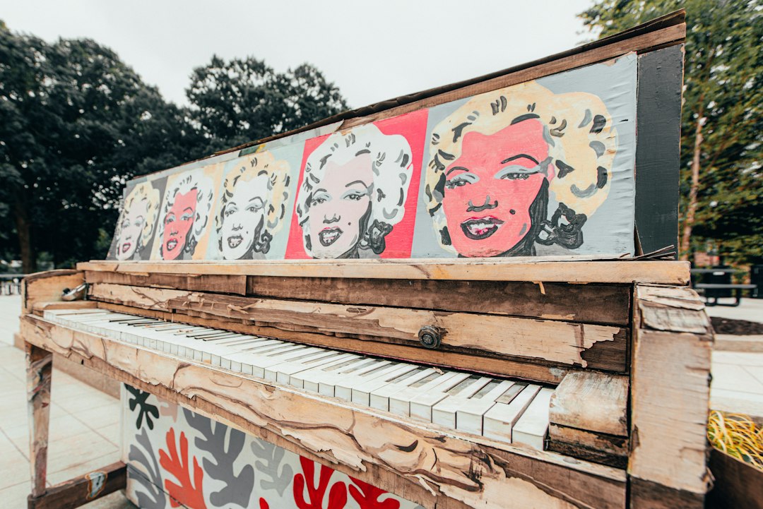 A weathered piano outdoors features pop art-style portraits of Marilyn Monroe, evoking the power of celebrity endorsements across its top panel. Trees and pavement are visible in the background.
