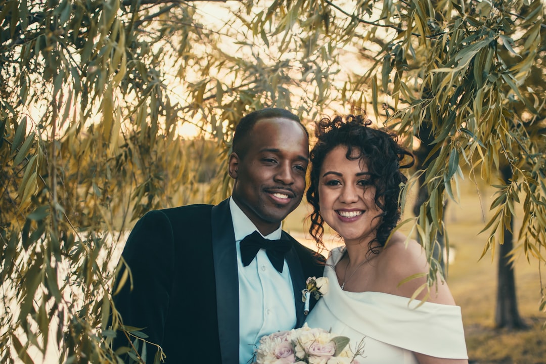 A couple dressed in formal wedding attire stands close together, smiling, surrounded by greenery and holding a bouquet, radiating happiness and flawless Bridal Beauty.