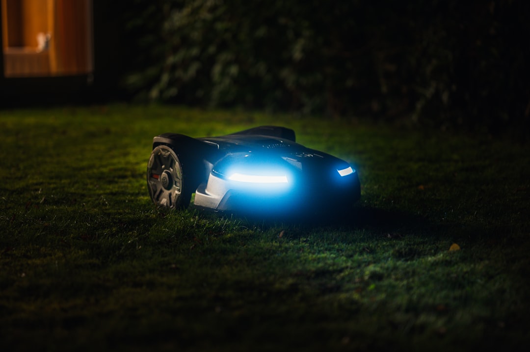 A robotic lawn mower with headlights operates on grass at night, its bright blue lights shining as if tending the lawn of a private island—perfect for those dreaming of Privatinseln zum Kaufen.