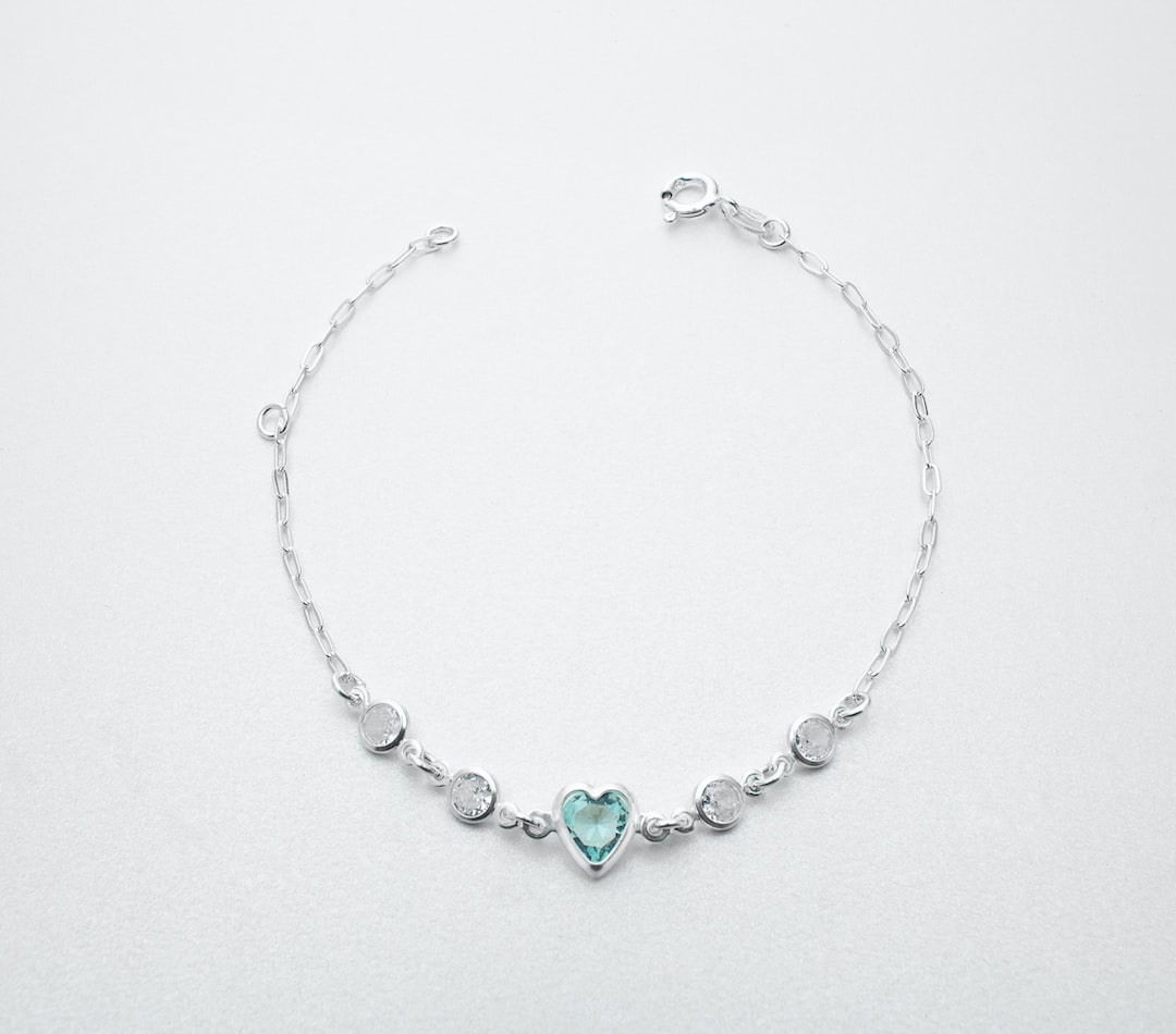 A silver chain bracelet featuring a central heart-shaped blue gemstone flanked by four round clear stones, this piece showcases elegance in jewelry design, displayed on a light gray background.