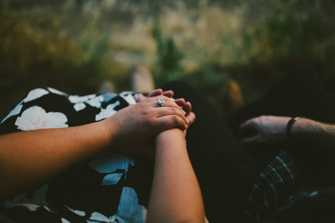 Close-up of two people holding hands, one wearing a floral dress and cultural jewelry—a sparkling engagement ring—celebrating beauty against a softly blurred outdoor background.