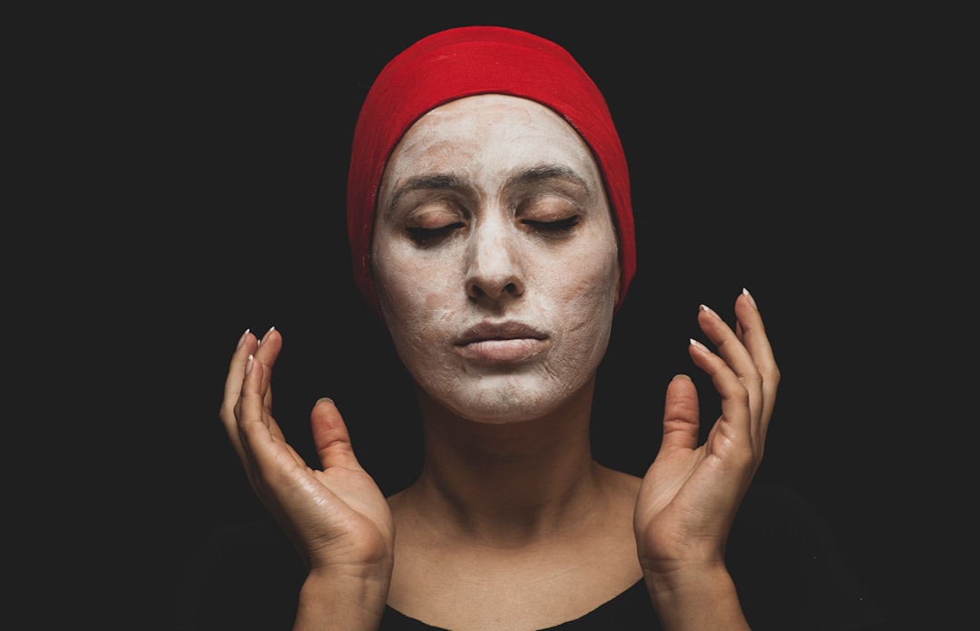 Person with eyes closed wearing a red head covering and a white facial mask, holding both hands near their face against a black background—perfect inspiration for sensitive skin makeup routines.