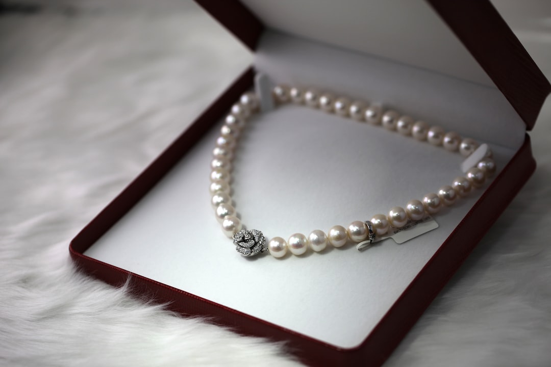 A pearl necklace with a decorative clasp, reflecting high-end jewelry craftsmanship, is displayed in an open red jewelry box on a white, fluffy surface.