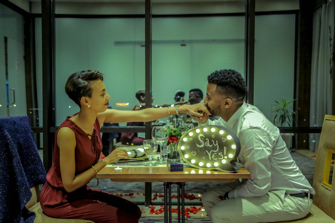 A couple sits at a table set for a romantic dinner, with a lit sign reading "Say Yes!" as the woman feeds the man, hinting at a marriage proposal and the excitement of choosing custom engagement rings.