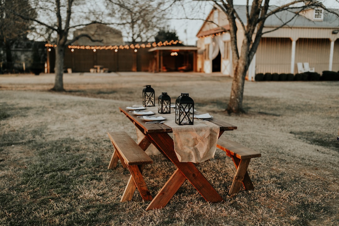 A wooden picnic table with benches is set for a meal outdoors, featuring black lanterns and a burlap runner, with a luxurious dining vibe reminiscent of Bangkok rooftop restaurants and string lights illuminating a charming barn backdrop.