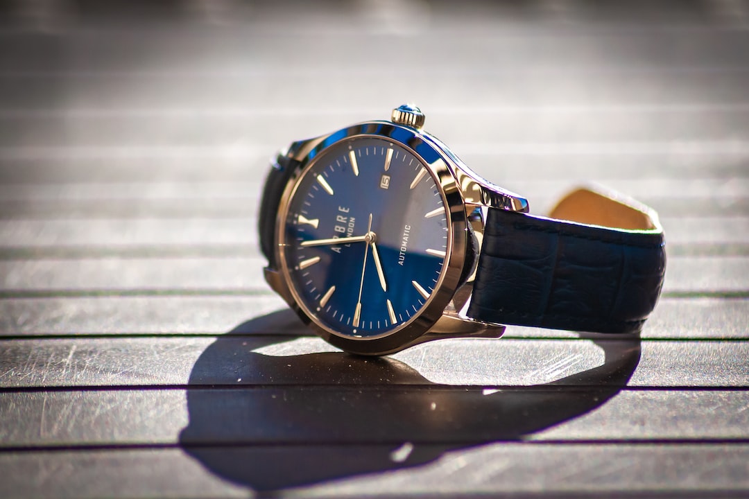 A close-up of a luxury watch with a blue face and leather strap, placed on a wooden surface with sunlight casting shadows, showcasing timeless style and personalization.