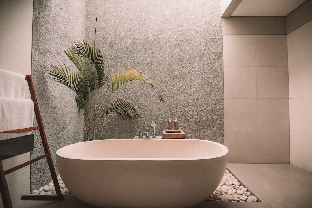 Modern bathroom with a freestanding tub, pebble floor accents, a green plant, wall-mounted faucet, and towel ladder against a gray textured wall—a serene space inspired by Danish design and luxury furniture homeware.