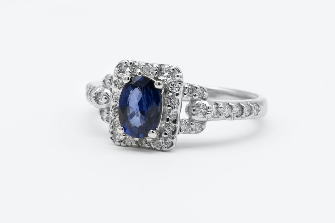 A silver ring with a rectangular setting featuring a central blue oval gemstone, surrounded by small clear stones and additional stones along the band, offers the perfect sparkle inspired by classic diamond rings.