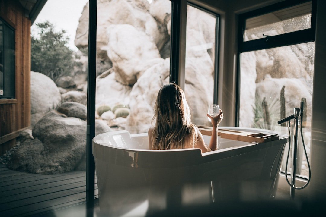 A person with long hair sits in a white bathtub, holding a glass and gazing out large windows at rocks and desert plants, creating the feel of a serene Desert Oasis.