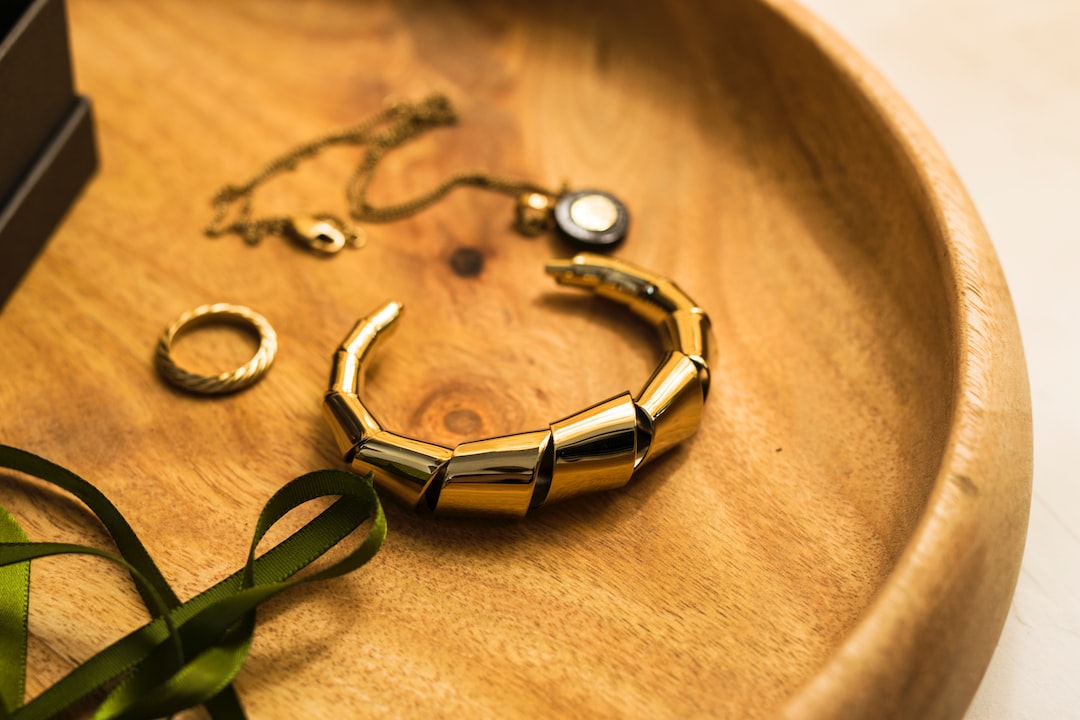 Gold jewelry, including a chunky bracelet, ring, and necklace with a black pendant, exudes elegance in a round wooden dish next to a green ribbon.