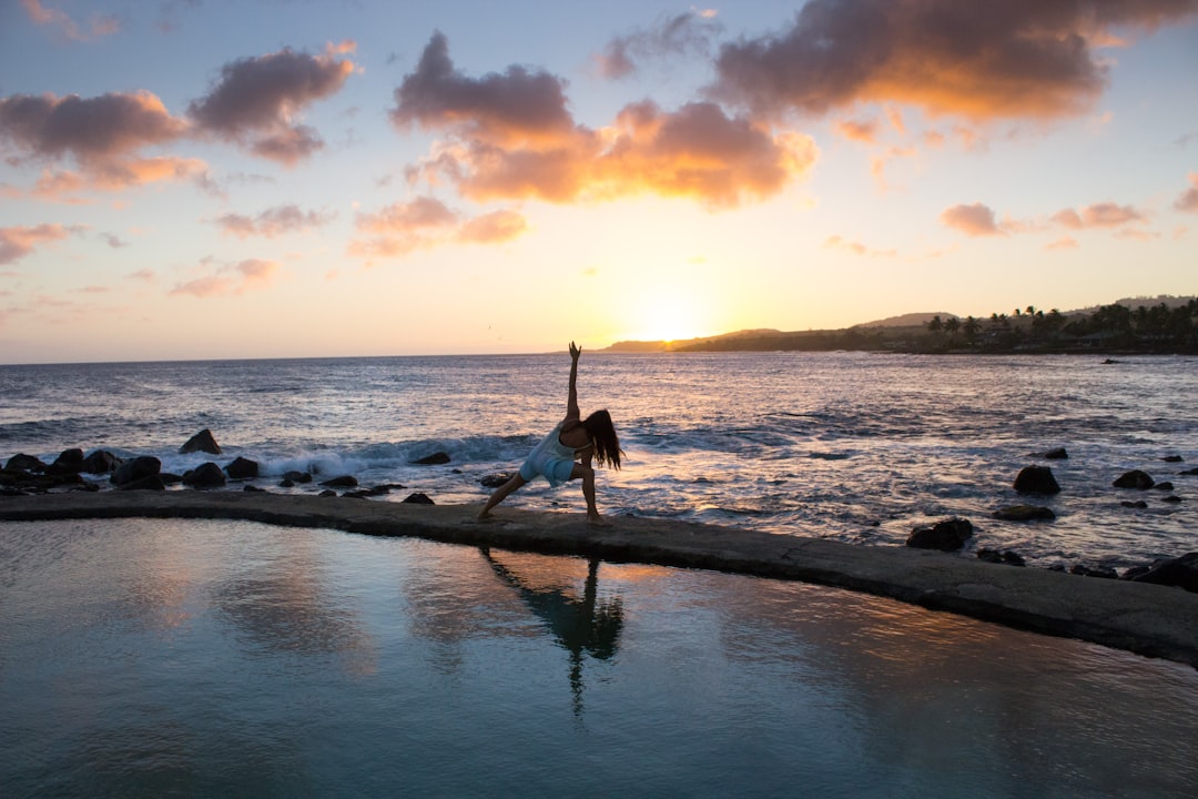 A person practices yoga near a calm pool of water at sunset, with the ocean and a partly cloudy sky in the background—embracing inner serenity found at exclusive destinations.