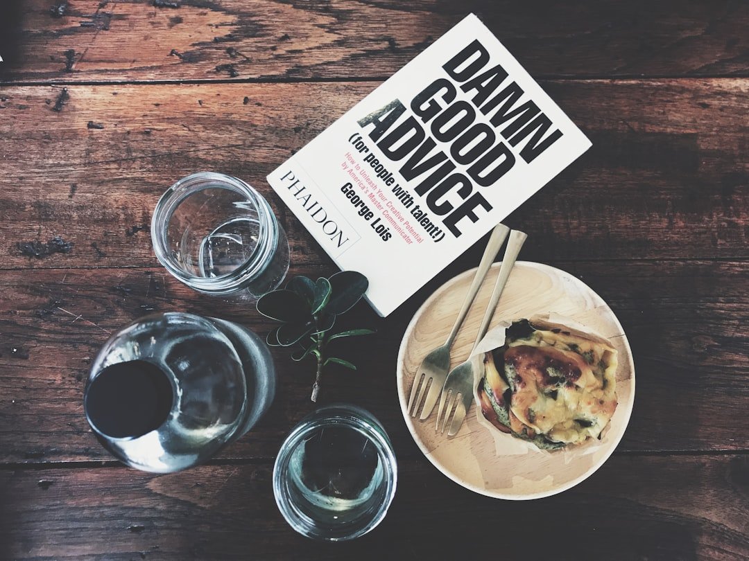 A book titled "Damn Good Advice" sits on a wooden table next to a plate of food, two glasses, a bottle, and a small plant—the perfect setup for planning your 2024 Top 10 luxury travel destinations.