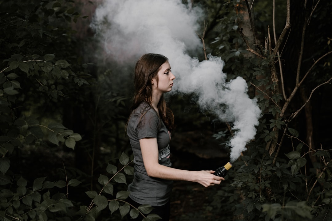 A woman stands in a forest in Africa holding a smoking device, releasing a thick cloud of white smoke that drifts to her left among the trees and foliage, capturing a moment of exotic bliss.