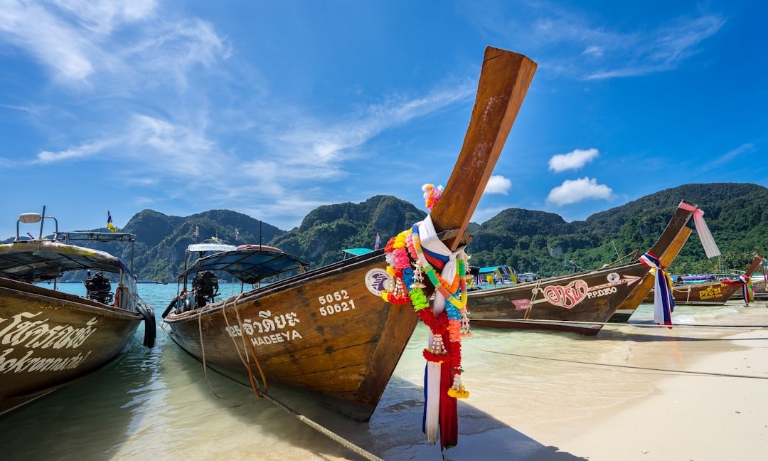 Wooden long-tail boats decorated with colorful ribbons and flowers are docked on a clear sandy beach in Thailand, with luxury beach resorts nestled among green hills and a blue sky in the background.