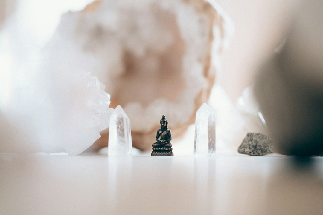 A small Buddha statue is placed on a white surface between clear quartz crystals, evoking the tranquil energy found at wellness escapes and retreats in Asia, with a blurred geode in the background.