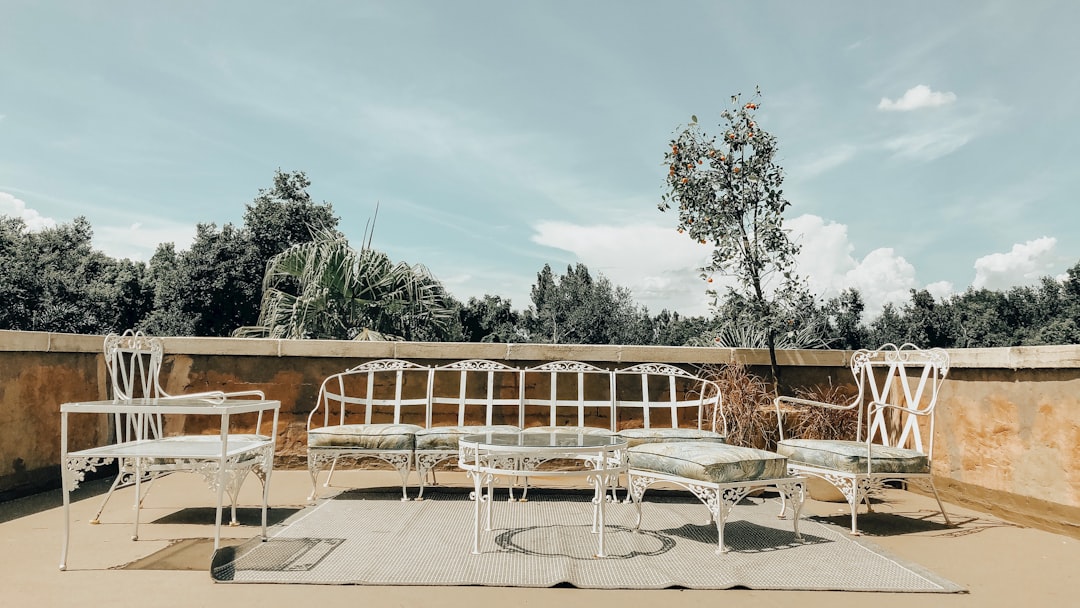 White metal patio furniture sits on a rug on an outdoor terrace, offering elevated elegance with a small tree and greenery in the background under a partly cloudy sky—reminiscent of exclusive rooftop bars.