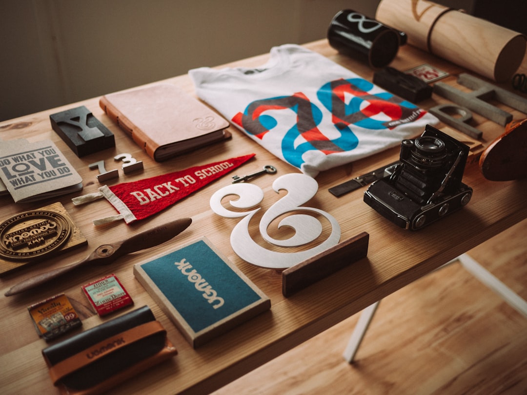 A wooden table displays assorted items, including a t-shirt, vintage camera, notebooks, a red pennant, a metal ampersand, stamps, and various small stationery objects—perfect inspiration for art and design hotels or exclusive travel destinations.