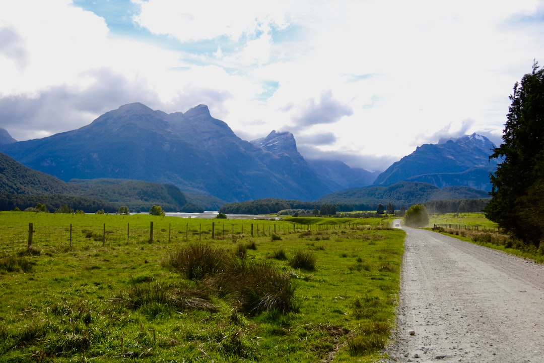 A gravel road runs through a green field with wire fencing, leading towards distant forested mountains under a cloudy sky—an inviting route to hidden paradises and secluded boutique hotels nestled beyond the horizon.