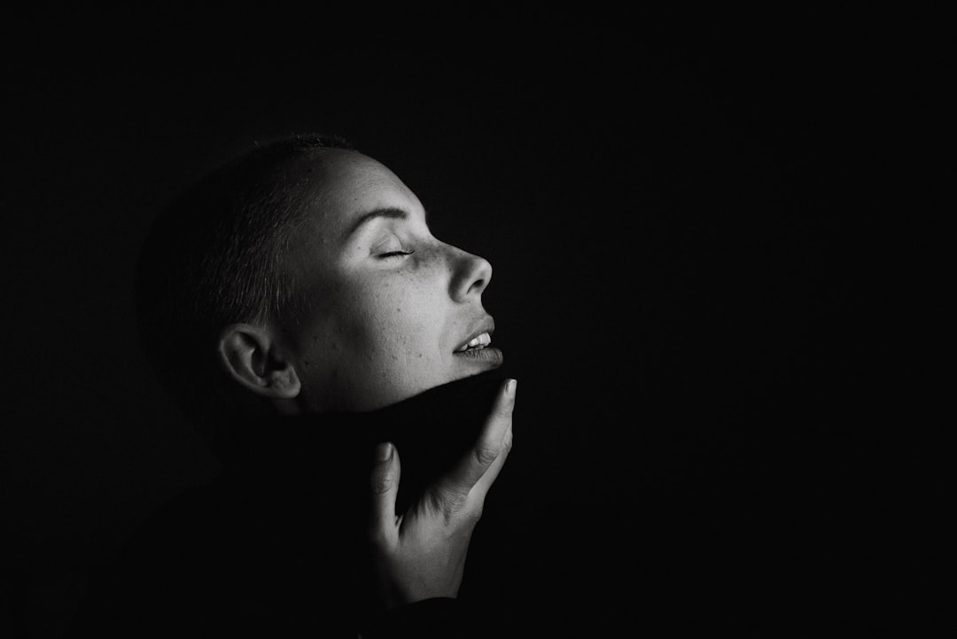 A person with closed eyes tilts their head back, holding their chin with one hand, illuminated by soft, dramatic lighting—capturing the calm confidence found in natural beauty and simple beauty solutions.