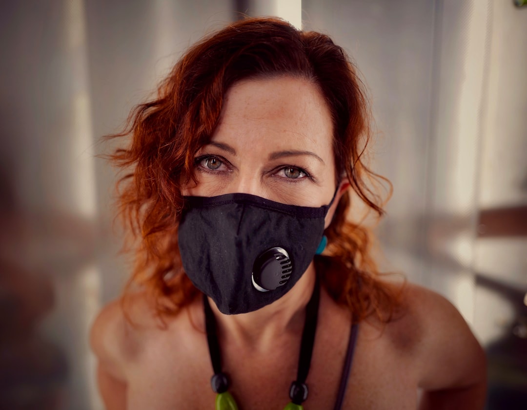 A woman with silky, wavy reddish hair wears a black face mask with a vent, a necklace with green beads, and stands indoors against a blurred background.