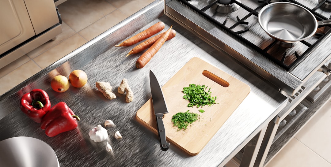 A stainless steel kitchen counter with carrots, apples, ginger, red bell pepper, garlic, chopped herbs on a cutting board, and a knife next to a gas stove with a pan—perfect for prepping natural lip balm or exploring DIY lip balm recipes.