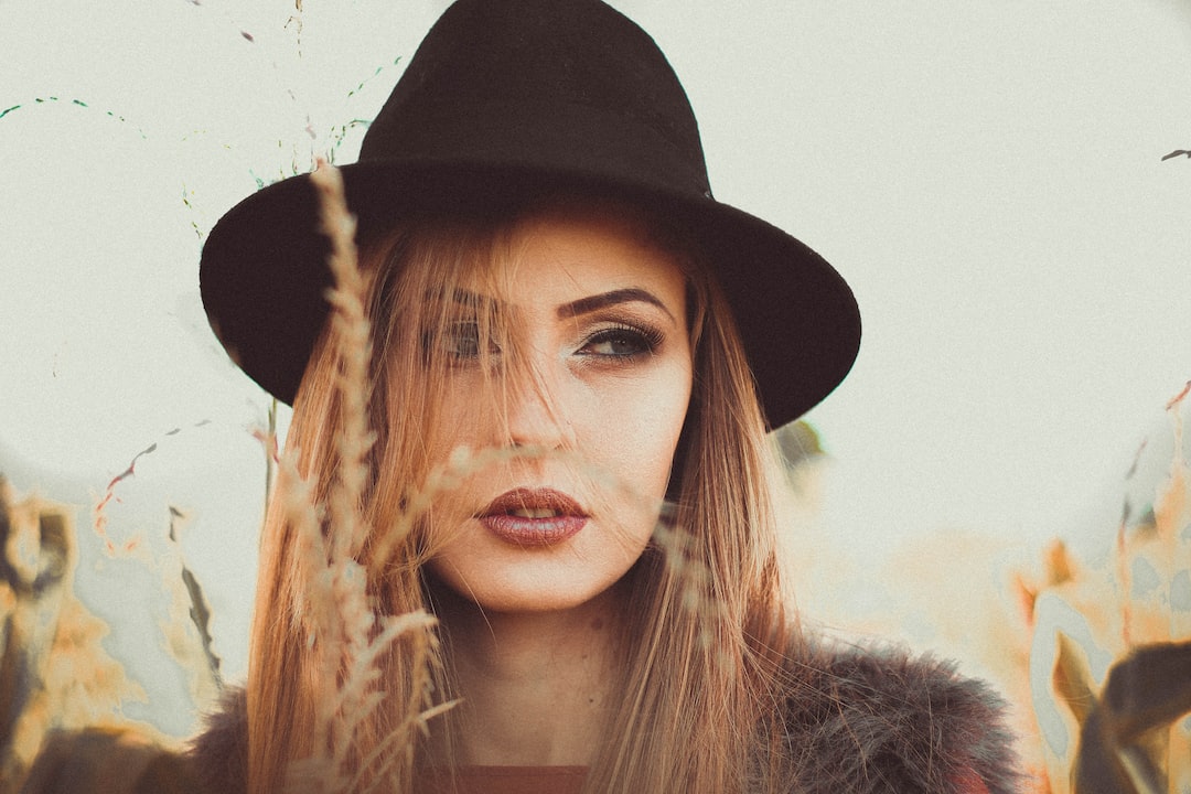 Woman with long hair wearing a black wide-brimmed hat, standing outdoors among tall grass, looking directly at the camera, exuding an artistic flair reminiscent of Dries Van Noten’s contemporary design sensibility.