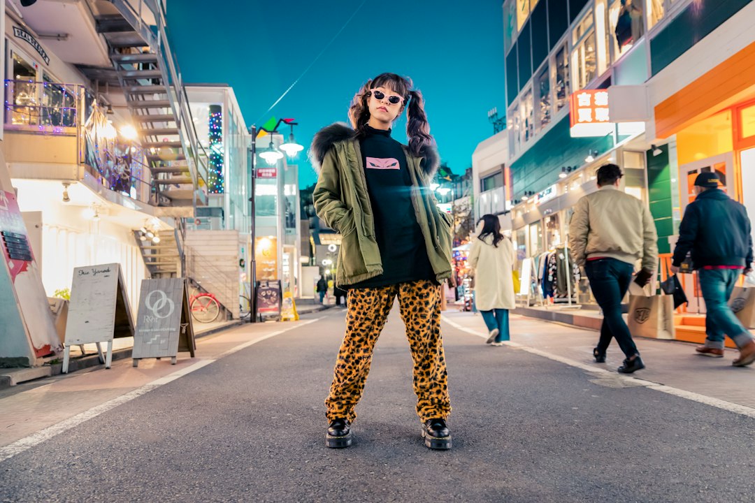 A person exudes sophistication, wearing sunglasses, a green jacket, and leopard print pants as they stand confidently in the middle of a brightly lit city street at night—an icon of women's fashion.