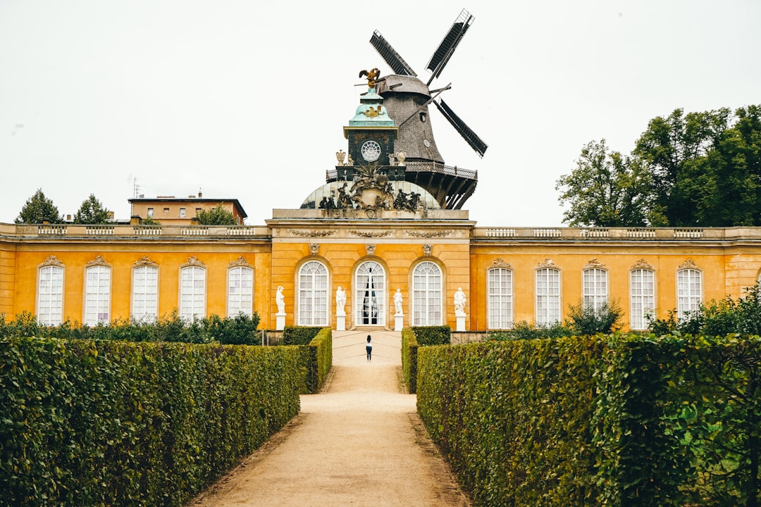 A yellow historic building with arched windows and statues in front, topped by a windmill, offers a unique paradise escape, seen from a gravel path lined with hedges.