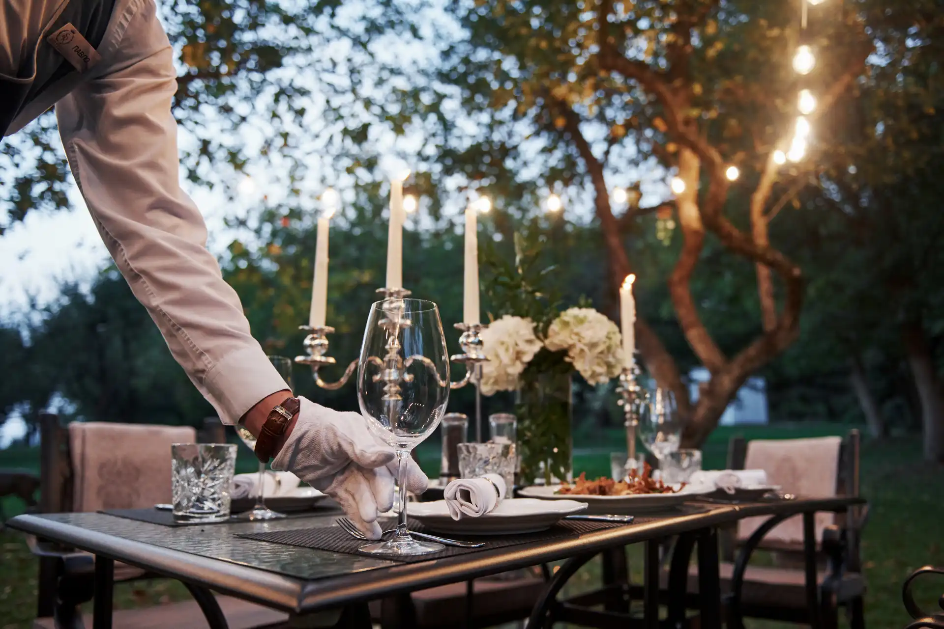 A gloved server sets a wine glass on a luxury outdoor dining table adorned with candles, flowers, and glowing string lights in the background.