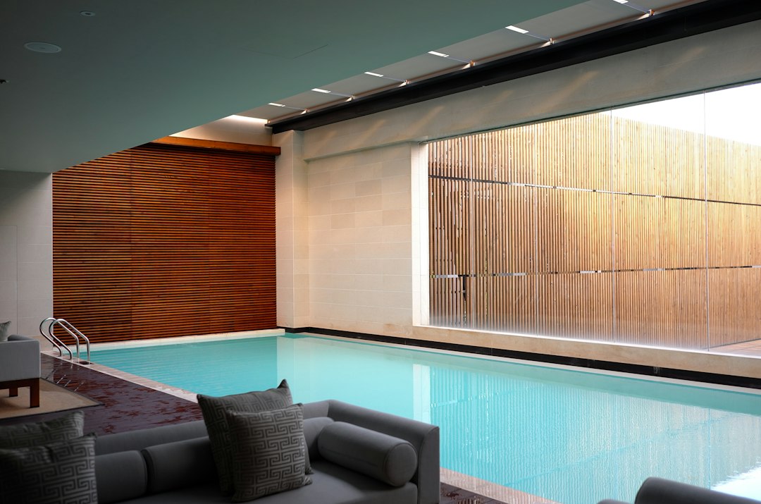 Modern indoor swimming pool with light blue water, beige walls, wooden slatted paneling, and a cushioned seating area in the foreground—perfect for relaxing at wellness resorts offering exclusive wellness experiences.