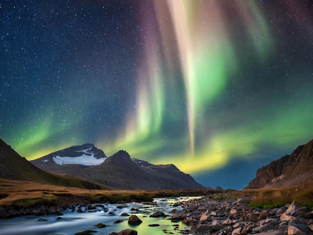 Colorful northern lights illuminate the night sky over a mountain wilderness, with a river and rocky terrain in the foreground—perfect for wildlife observations.