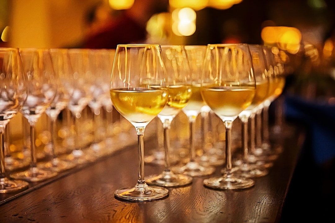Several glasses of white wine are arranged in rows on a wooden surface, with warm ambient lighting in the background—perfect for exclusive wine tasting experiences.