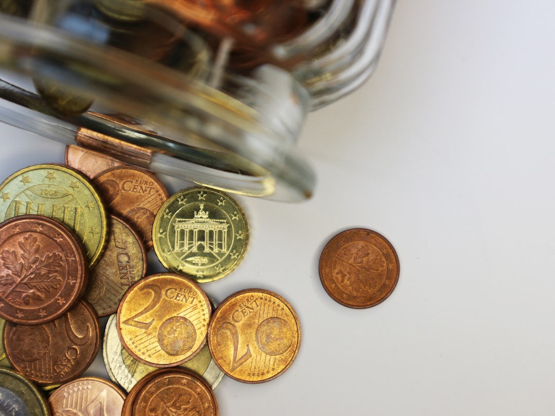A glass jar tipped over with various euro cent coins spilling onto a white surface suggests the start of a Europe travel adventure; one coin is separate from the group.