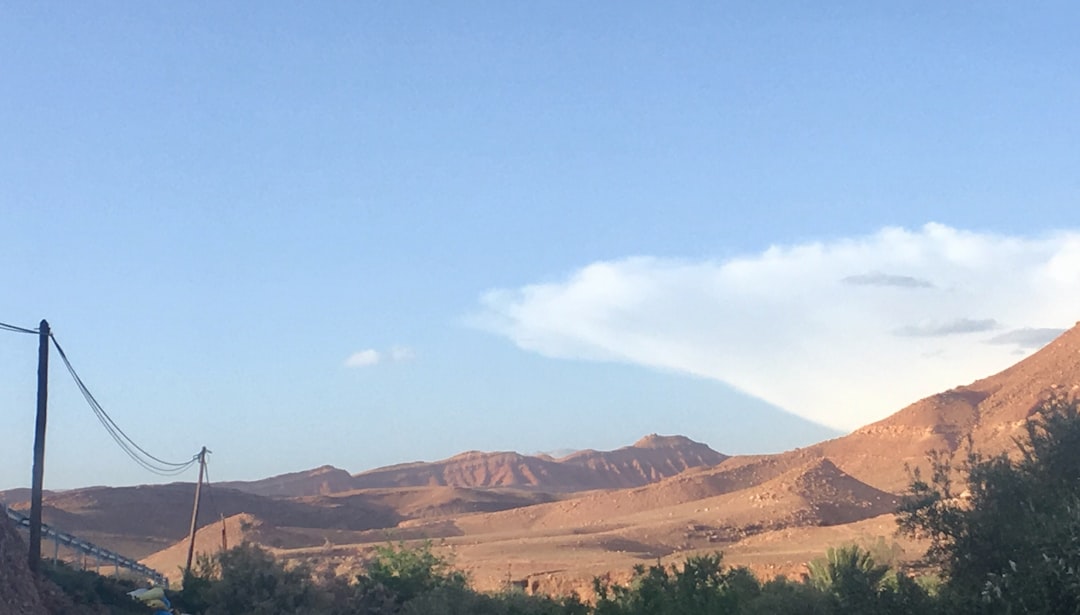 A desert landscape in Morocco with brown hills, sparse vegetation, power lines, and a clear blue sky dotted with white clouds—perfect for those seeking a desert adventure.
